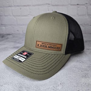 May include: A green and black trucker hat with a brown leather patch that says "Mike & Mandy X.XXXIII.MMXXXIII".