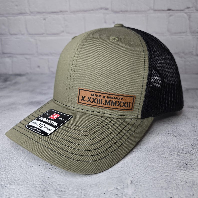 May include: A green and black trucker hat with a brown leather patch that says "Mike & Mandy X.XXXIII.MMXXXIII".