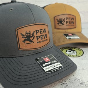 May include: Two trucker hats. One is gray with a black mesh back and a brown leather patch featuring a cartoon cat with guns and the words "PEW PEW MADAFAKASI." The other is tan with a similar design. Both have "RICHARDSON" tags.