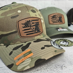 May include: Camouflage baseball cap with a brown leather patch featuring a bow hunter silhouette and American flag design. The cap has a black mesh back and a curved brim. The official licensed product sticker is visible on the brim.