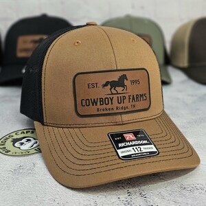 K&ouml;nnte beinhalten: Braune Trucker-Cap mit schwarzem Mesh-R&uuml;cken. Sie hat einen Lederaufn&auml;her mit einer Pferdesilhouette und dem Text "COWBOY UP FARMS" und "Broken Ridge, TN". Die Kappe ist verstellbar und tr&auml;gt das Markenetikett "RICHARDSON".