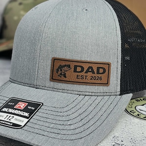 May include: A gray trucker hat with a black mesh back. The hat features a brown leather patch with a fish graphic and the text "DAD EST. 2026". The Richardson brand tag is visible on the brim.