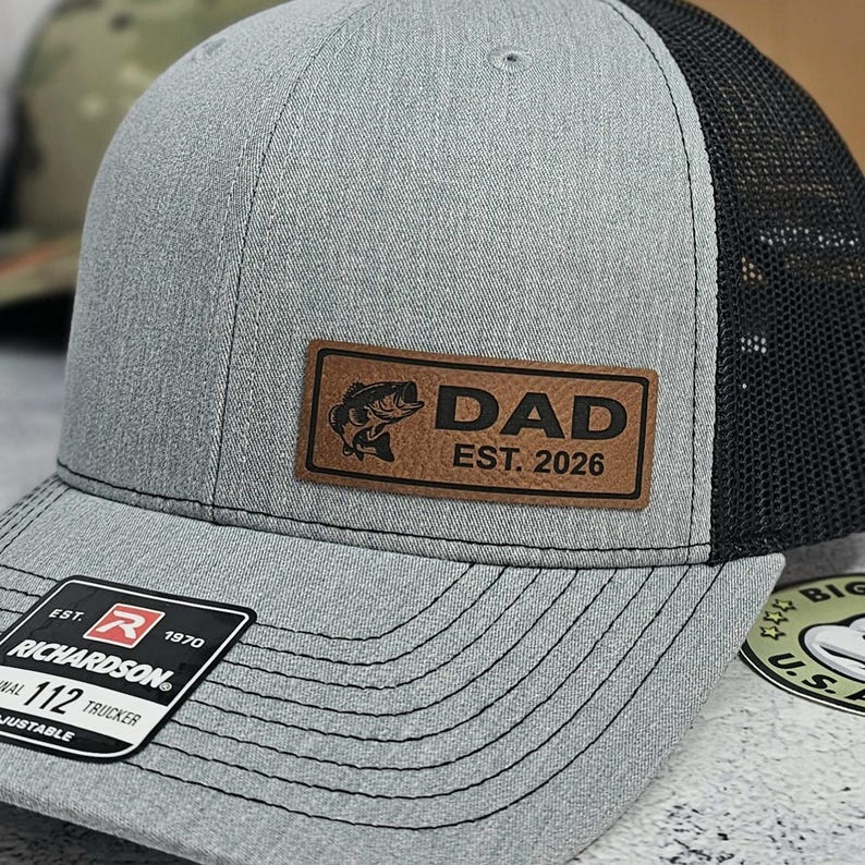 May include: A gray trucker hat with a black mesh back. The hat features a brown leather patch with a fish graphic and the text "DAD EST. 2026". The Richardson brand tag is visible on the brim.