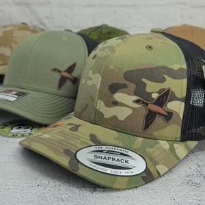 May include: A collection of snapback trucker hats with a duck logo. The hats feature a variety of camouflage patterns, including green, brown, and tan. The hats have a mesh back and a curved brim. The hats have a "Snapback" label.