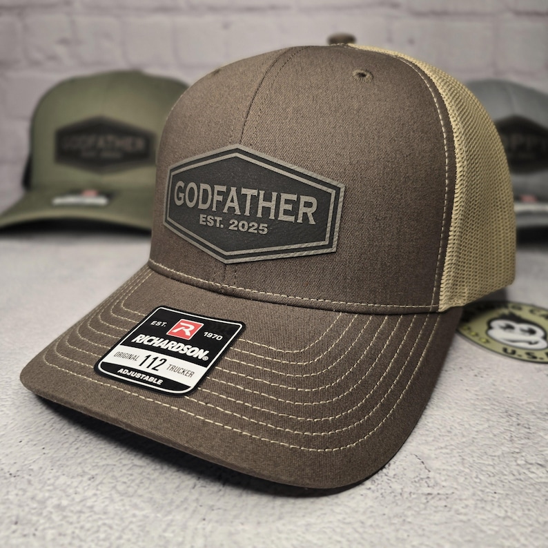 Puede incluir: Un gorro de camionero marr&oacute;n y beige con un parche de cuero negro que dice "GODFATHER EST. 2025". El gorro est&aacute; hecho por Richardson 112.