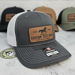 K&ouml;nnte beinhalten: Graue Trucker-Cap mit wei&szlig;em Mesh-R&uuml;cken und braunem Lederpatch. Der Patch zeigt eine Pferdesilhouette, den Text "EST. 1995" und Platz f&uuml;r individuellen Text. Die Kappe ist verstellbar und hat ein Richardson-Etikett.