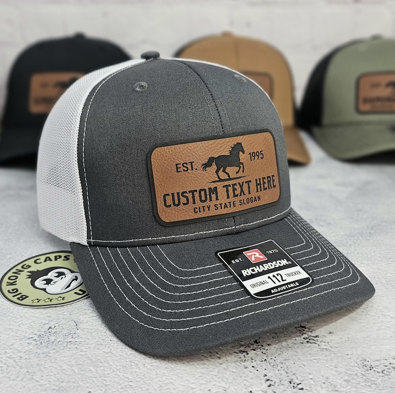 K&ouml;nnte beinhalten: Graue Trucker-Cap mit wei&szlig;em Mesh-R&uuml;cken und braunem Lederpatch. Der Patch zeigt eine Pferdesilhouette, den Text "EST. 1995" und Platz f&uuml;r individuellen Text. Die Kappe ist verstellbar und hat ein Richardson-Etikett.