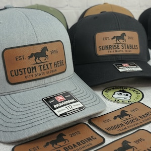 K&ouml;nnte beinhalten: Graue Trucker-Cap mit einem braunen Lederaufn&auml;her mit einer Pferdesilhouette und dem Text "CUSTOM TEXT HERE". Die Kappe hat eine schwarze Mesh-R&uuml;ckseite und ein Richardson 112-Etikett. Weitere Kappen mit Pferdelogos sind sichtbar.