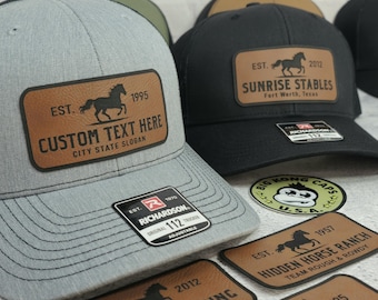 May include: Gray trucker hat with a brown leather patch featuring a horse silhouette and the text "CUSTOM TEXT HERE". The hat has a black mesh back and a Richardson 112 tag. Other hats with horse logos are visible.