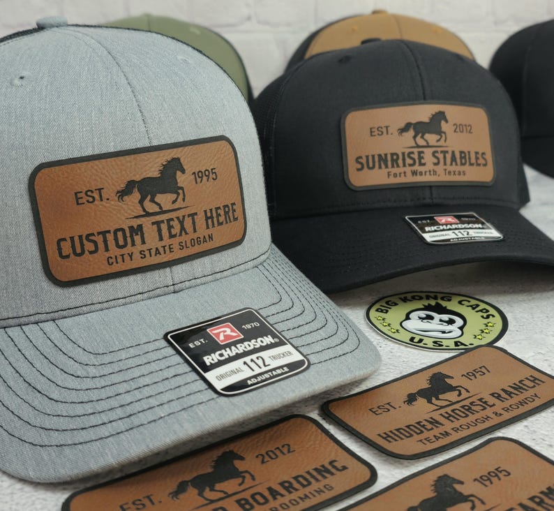 K&ouml;nnte beinhalten: Graue Trucker-Cap mit einem braunen Lederaufn&auml;her mit einer Pferdesilhouette und dem Text "CUSTOM TEXT HERE". Die Kappe hat eine schwarze Mesh-R&uuml;ckseite und ein Richardson 112-Etikett. Weitere Kappen mit Pferdelogos sind sichtbar.