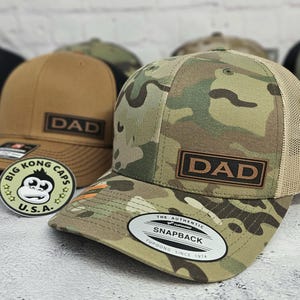 May include: Camouflage trucker hat with a brown leather patch that reads "DAD". The hat has a curved brim and a mesh back. A second brown hat with "DAD" patch is visible in the background. A "Big Kong Caps U.S.A." sticker is in the foreground.