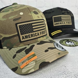 May include: Two camouflage baseball caps with the American flag and the text "AMERICA FIRST". One cap has a black mesh back. The other cap is black with a tan flag. A sticker on the front of the cap reads "MULTICAM".