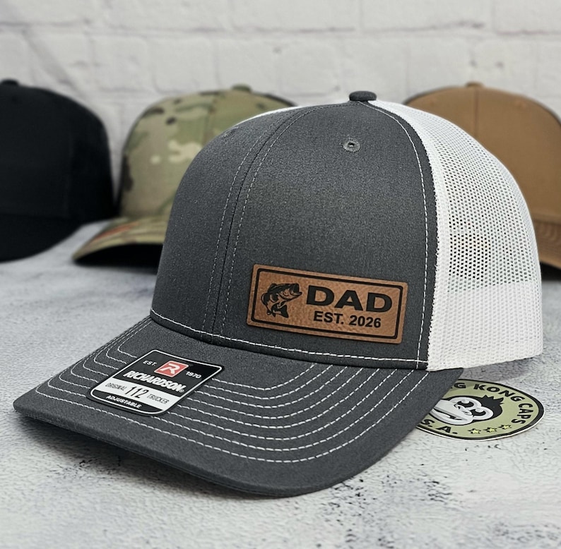 May include: A gray baseball cap with a white mesh back and a brown leather patch that reads "DAD EST. 2026" with a fish graphic. The cap has a curved brim and white stitching. Other caps are visible in the background.
