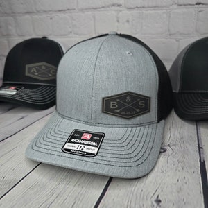 May include: A gray and black trucker hat with a black leather patch that says "B & S 08.24.25" on the front. The hat is adjustable and has a red and black Richardson label on the back.