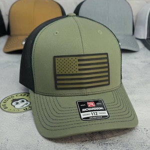 May include: An olive green trucker hat with a black mesh back and a rectangular patch featuring the American flag. The hat has a curved brim and an adjustable strap. The brand name "Richardson" is visible on a tag.
