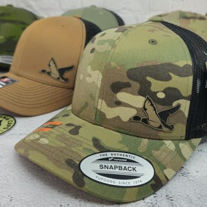 May include: A collection of baseball caps with a duck logo. One cap features a camouflage pattern, black mesh back, and a "Snapback" label. Other caps are tan and olive green. The caps have a curved brim and adjustable snap closure.