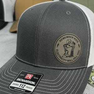 May include: A gray trucker hat with a white mesh back and a curved brim. The hat features a circular patch with the text "REMEMBER KIDS ELECTRICITY WILL KILL YOU" and a cartoon illustration. The Richardson brand tag is visible.