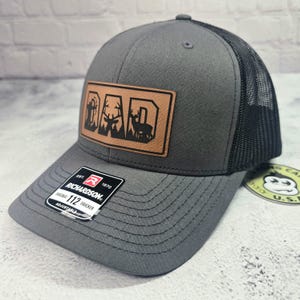 May include: Gray trucker hat with a black mesh back and a brown leather patch. The patch features the word "DAD" with a silhouette of a deer and trees. The hat has a curved brim and an adjustable strap.