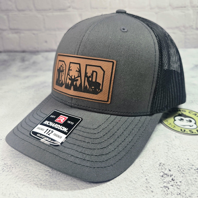 May include: Gray trucker hat with a black mesh back and a brown leather patch. The patch features the word "DAD" with a silhouette of a deer and trees. The hat has a curved brim and an adjustable strap.
