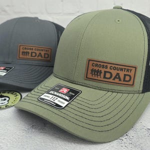 May include: Two trucker hats with a Richardson patch. One hat is olive green with a black mesh back and a brown leather patch that reads "CROSS COUNTRY DAD". The other hat is gray with a black mesh back and a similar patch.