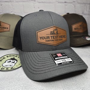 May include: A gray and black trucker hat with a brown leather patch that says "YOUR TEXT HERE CUSTOM TEXT HERE". The patch features a small illustration of an excavator. The hat is adjustable and has a black mesh back.