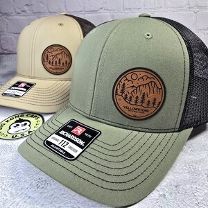 May include: Two trucker hats with a circular patch featuring a mountain and tree design. One hat is tan with a brown mesh back, the other is olive green with a black mesh back. Both hats have the text "YELLOWSTONE NATIONAL PARK" on the patch.