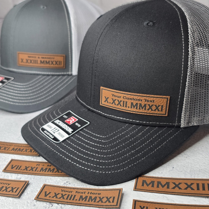 May include: Two black and gray trucker hats with a brown leather patch on the front. The patch has the text "Your Custom Text X.XXII.MMXXI" on it. There are several brown leather patches with Roman numeral dates on a gray surface.