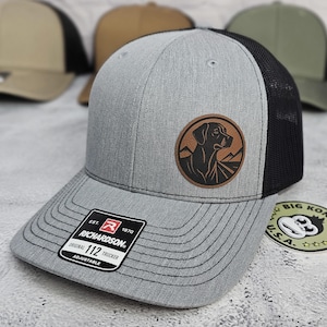 May include: A gray Richardson 112 trucker hat with a black mesh back and a brown leather patch featuring a dog silhouette and mountain graphic. The hat has a curved brim and an adjustable strap.