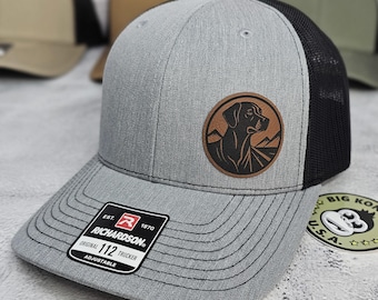 Könnte beinhalten: Graue Richardson 112 Trucker-Kappe mit schwarzem Mesh-Rücken und einem braunen Lederaufnäher mit einer Hunde-Silhouette und einem Berg-Motiv. Die Kappe hat einen gebogenen Schirm und einen verstellbaren Riemen.