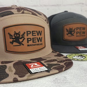 May include: Two flat bill snapback hats. One hat is camouflage with a brown leather patch featuring a black cat holding guns and the words "PEW PEW MADAFKASI". The other hat is dark gray with a similar patch.