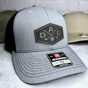 May include: Gray trucker hat with a black mesh back and a curved brim. The hat features a leather patch with the word "DAD" and golf clubs. The Richardson 112 hat is adjustable.