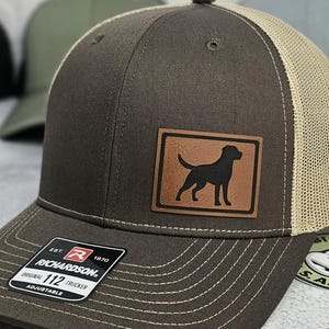 May include: A brown trucker hat with a tan mesh back and a brown leather patch featuring a black dog silhouette. The hat has a curved brim and the Richardson 112 Trucker label.