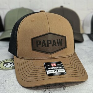 May include: A brown trucker hat with a black mesh back and a curved brim. The hat features a leather patch with the word "PAPAW" and a Richardson 112 tag. Adjustable fit.