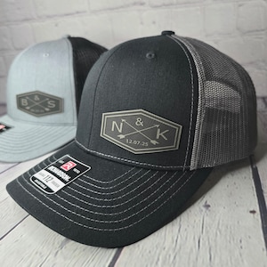 May include: Two black and gray trucker hats with embroidered patches. The first hat has a patch with the letters "B & S" and crossed arrows. The second hat has a patch with the letters "N & K" and crossed arrows, and the date "12.07.25".