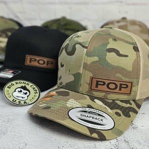 May include: A camouflage print Snapback hat with a tan mesh back and a brown leather patch that reads "POP". A black hat with a brown leather patch that reads "POP" is behind it. A sticker with a monkey graphic is in the foreground.