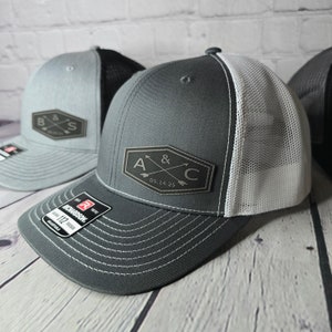 May include: Two gray and white trucker hats with black leather patches. The patches feature a monogram with two initials and an ampersand, along with a date. One patch reads "A & C 05.14.25" and the other reads "B & S".