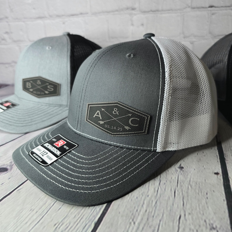 May include: Two gray and white trucker hats with black leather patches. The patches feature a monogram with two initials and an ampersand, along with a date. One patch reads "A & C 05.14.25" and the other reads "B & S".