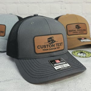 May include: A gray trucker hat with a black mesh back and a brown leather patch. The patch features a truck graphic and the text "CUSTOM TEXT YOUR CUSTOM TEXT". The hat is adjustable and has a Richardson 112 Trucker tag.
