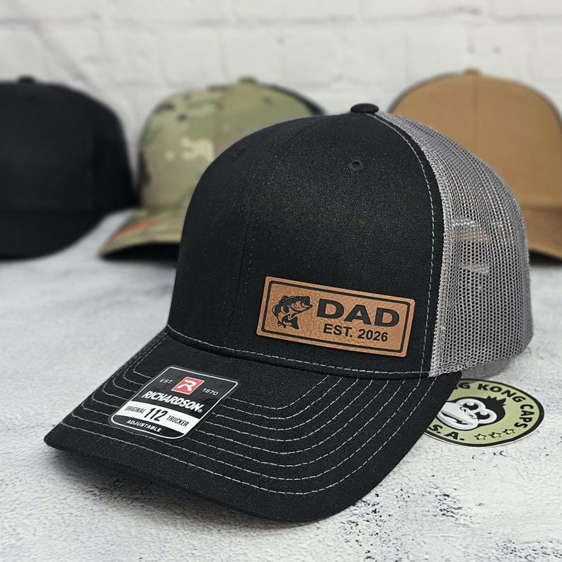 May include: Black Richardson 112 trucker hat with a gray mesh back. The hat features a brown leather patch with the word "DAD", a fish graphic, and "EST. 2026". The brim is curved with white stitching. The hat is adjustable.