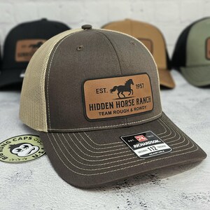 K&ouml;nnte beinhalten: Braune Trucker-Cap mit beigem Mesh-R&uuml;cken und Lederaufn&auml;her. Der Aufn&auml;her zeigt eine Pferdesilhouette und den Text "HIDDEN HORSE RANCH". Die Kappe ist verstellbar und hat einen gebogenen Schirm.