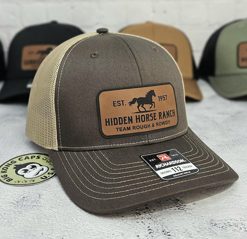 K&ouml;nnte beinhalten: Braune Trucker-Cap mit beigem Mesh-R&uuml;cken und Lederaufn&auml;her. Der Aufn&auml;her zeigt eine Pferdesilhouette und den Text "HIDDEN HORSE RANCH". Die Kappe ist verstellbar und hat einen gebogenen Schirm.