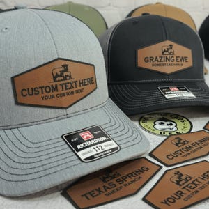 May include: A collection of trucker hats with custom leather patches. One gray hat features a patch with the text "CUSTOM TEXT HERE" and a sheep graphic. A black hat displays "GRAZING EWE" and "HOMESTEAD HAVEN".