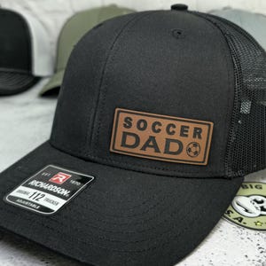 May include: Black trucker hat with a mesh back and a brown leather patch that reads "SOCCER DAD" with a soccer ball graphic. The hat has a curved brim and an adjustable strap.