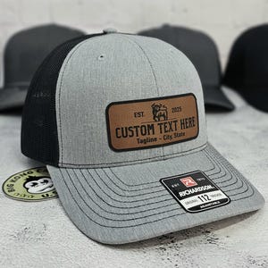 May include: A gray trucker hat with a black mesh back and a brown leather patch. The patch reads "CUSTOM TEXT HERE" with a bull graphic and the text "Tagline - City, State". The hat has a Richardson 112 Trucker tag.