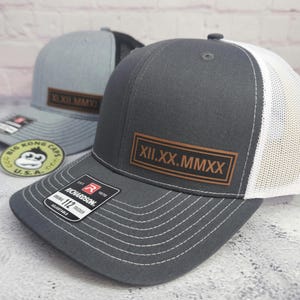 May include: Two trucker hats. One is gray with a white mesh back and a brown leather patch with Roman numerals. The other is gray with a black mesh back and a brown leather patch with Roman numerals. The hats have a Richardson tag.