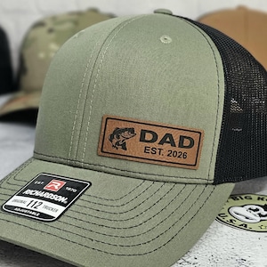 May include: Olive green trucker hat with a black mesh back. A brown leather patch features a fish graphic and the word "DAD" with "EST. 2026". The hat has a curved brim and an adjustable strap.