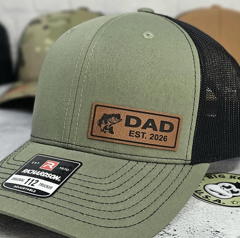 May include: Olive green trucker hat with a black mesh back. A brown leather patch features a fish graphic and the word "DAD" with "EST. 2026". The hat has a curved brim and an adjustable strap.