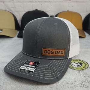 May include: A gray trucker hat with a white mesh back and a curved brim. The hat features a brown leather patch with the words "DOG DAD" and a Richardson label. Other hats are visible in the background.