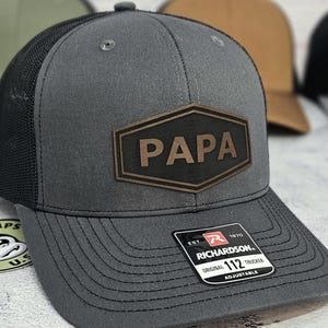 May include: Gray trucker hat with a black mesh back and a brown leather patch that reads "PAPA". The hat has a curved brim and an adjustable strap. The Richardson 112 Trucker hat is a classic style.