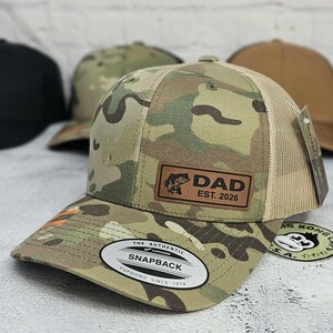 May include: Camouflage print Snapback hat with a brown leather patch that reads "DAD EST. 2026" and a fish graphic. The hat has a tan mesh back and a curved brim. The hat has a white tag that reads "THE AUTHENTIC SNAPBACK YUPOONG SINCE 1974".